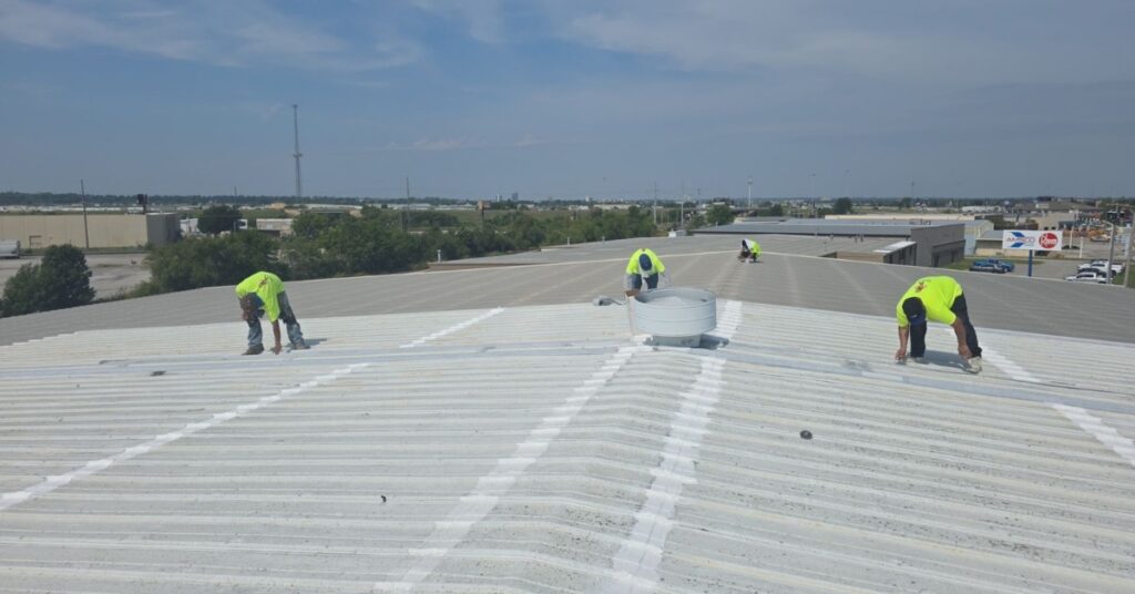 Three construction workers in neon shirts kneeling on a white corrugated metal roof, performing maintenance on a sunny day.