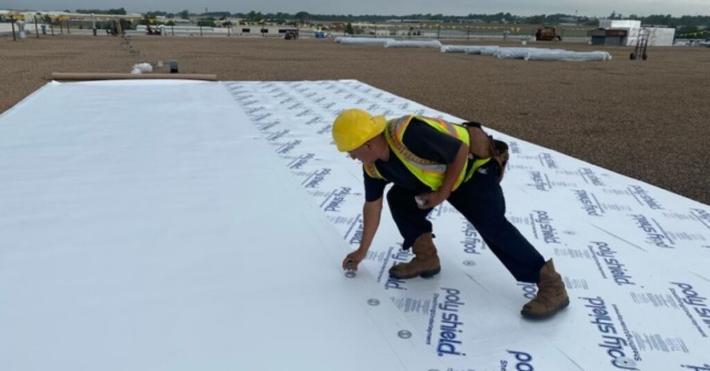 Construction worker in a yellow hard hat and high‑visibility vest kneels on a white roof underlayment, securing panels.