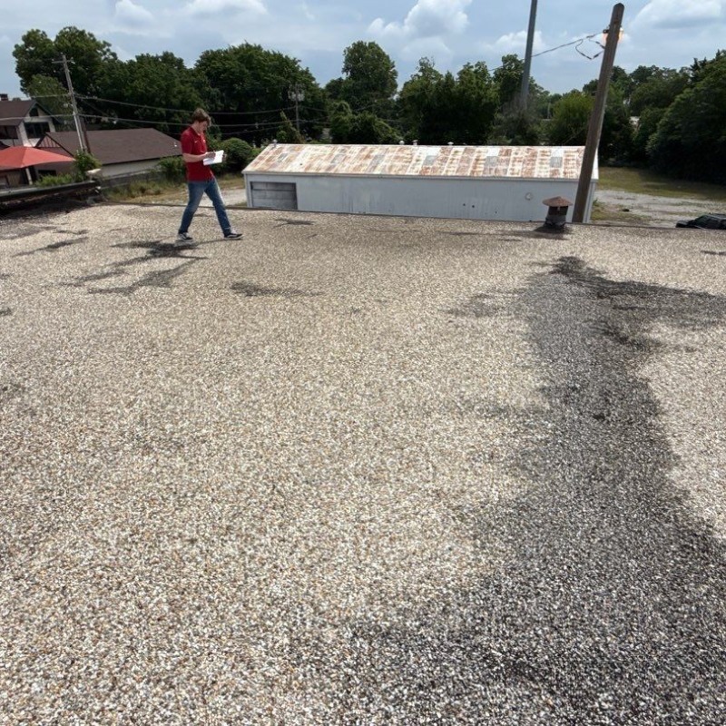 Person in a red shirt walks across a flat gravel rooftop, with a small shed and power lines in the background.