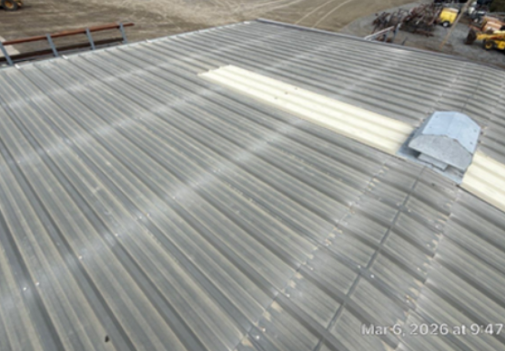 Wide view of a corrugated metal roof under construction, with a white rectangular skylight patch and a small vent near the ridge; construction equipment visible in the distance and a timestamp reads Mar 6, 2026 at 9:47.